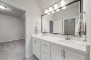 Modern bathroom interior with a dual-sink white vanity, large mirror, and carpeted hallway in the background.