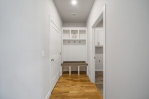 A bright, narrow hallway featuring a wooden bench under a row of hooks and cabinets, with white walls and hardwood flooring.