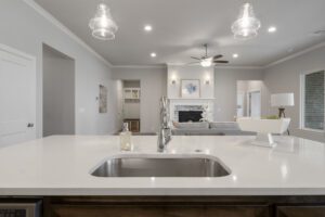 Modern kitchen with white countertops, sink, and stainless steel faucet, looking into an open-plan living area with elegant light fixtures.