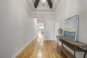 A bright, modern hallway with hardwood floors, white walls, beamed ceilings, a console table, and a large painting on the left.