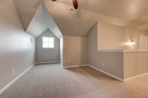 Interior of an empty attic room with slanted ceilings, two windows, and gray carpeting.