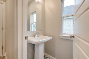 A small, bright bathroom featuring a pedestal sink, a mirror, and a window with a view of neighboring houses.