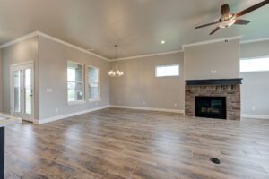 A spacious living room with gray walls, hardwood floors, a fireplace, ceiling fan, and large windows letting in natural light.