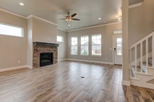 Spacious living room featuring a brick fireplace, wooden flooring, large windows, and a ceiling fan. neutral color tones with staircase on the side.