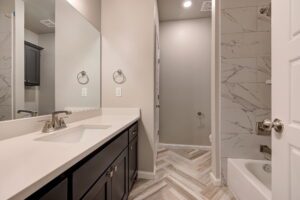 Modern bathroom with double vanity, dark cabinets, white countertops, and herringbone tile floor leading to a walk-in shower.