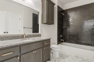 Modern bathroom with gray cabinetry, white countertop, hexagonal tile floor, and a bathtub enclosed by dark glass tiles.