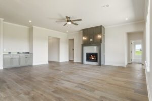 Spacious, empty living room featuring hardwood floors, gray kitchen cabinets, lit fireplace, and ceiling fan.