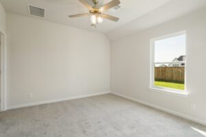 Empty room with beige carpet, white walls, a ceiling fan, and a large window showing a view of a fenced backyard.
