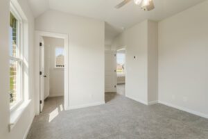 Empty new residential room with carpeted floor, white walls, windows, and a ceiling fan, leading to another room with a visible door.