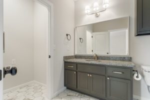 Modern bathroom interior featuring gray cabinets, granite countertops, white walls, and a large mirror with overhead lighting.