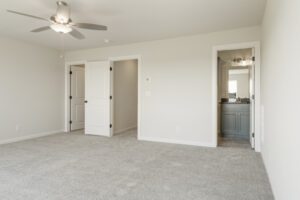 Empty room with beige carpet, white walls, a ceiling fan, and an open doorway leading to a kitchen.