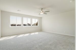 An empty, bright room with a gray carpet, white walls, a ceiling fan, and a three-panel window showing a suburban view.