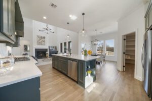 Modern kitchen with an island, stainless steel appliances, pendant lighting, and hardwood floors leading to an adjoining room with a fireplace.