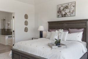 A serene and elegantly appointed bedroom featuring a dark wood bed frame with matching side table and lamp, crisp white bedding with accent pillows, decorative wall art above the bed, and a view into an en-suite bathroom.