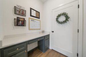 A small office nook with a built-in desk, green cabinets, and a white wall decorated with a wreath and framed items.