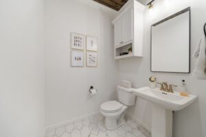 A clean and minimalistic bathroom with white walls, a pedestal sink, a framed mirror, a toilet, and decorative wall art.