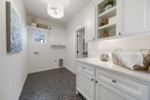 Modern laundry room with white cabinetry, herringbone tile floor, decorative items on shelves, and a painting on the wall.