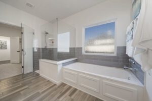 Modern bathroom with a freestanding tub, glass shower stall, gray tiles, wooden floor, and frosted window.