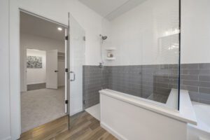 Modern bathroom with a glass shower, gray herringbone tiles, and white walls, featuring an adjacent room visible through an open door.