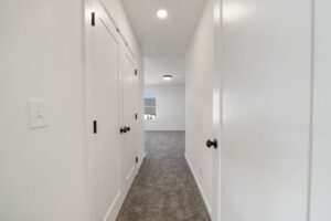 A narrow hallway in a modern home with white walls, carpeted floor, and several closed doors with black handles.