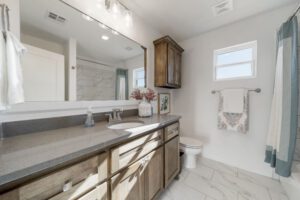 Modern bathroom with a large mirror, wooden cabinets, gray countertop, and white tiled flooring. towels hang beside the toilet and window.