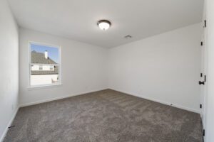 Empty, carpeted room with white walls and a large window showing a suburban view.