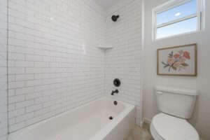 Modern, clean bathroom with white subway tiles, a bathtub with black fixtures, a toilet, and a floral painting above it. there's a small window allowing natural light.