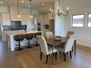 A modern kitchen and dining area with a wooden table, four chairs, island with three barstools, pendant lights, and a chandelier. White cabinets and light wood flooring complete the space.