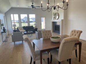 A modern living and dining room with a wooden table and cushioned chairs, a gray sofa set near the fireplace, large windows, and light wood flooring.