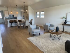 Open-plan living space with a kitchen, dining area, and sitting area. Features hardwood floors, pendant lighting, a dining table, and upholstered chairs. Neutral tones dominate the decor.