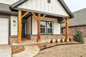 A modern house exterior featuring a front porch with wooden columns, a gray door, and three windows. The yard has minimal landscaping with small shrubs and mulch.