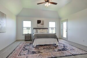 A bedroom with a bed, two windows, and a ceiling fan. The room features a rug, a nightstand with a lamp, and framed artwork on the walls. Natural light fills the space.