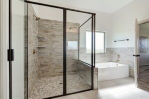 A modern bathroom features a glass-enclosed shower with stone tile flooring, a separate bathtub, a window, and white walls.