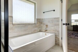 Bathroom with a modern white bathtub, wood-patterned tile walls, frosted window, and an empty towel rack. An open door leads to a carpeted room with a window.