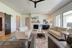 Modern living room with two beige sofas, a fireplace, a patterned rug, and large windows. Black sliding door on the side.