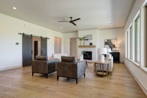 A modern living room with two gray armchairs, a white sofa, a wooden coffee table, and a fireplace. A ceiling fan is above, and large windows line the right wall.