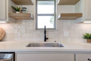 A kitchen sink under a window, surrounded by white cabinets and wooden shelves, with a potted plant and kitchen items displayed.