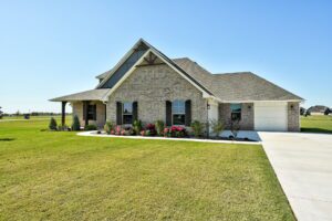 Brick house with a front porch, three windows, and a neatly maintained lawn. A driveway leads to a garage on the right.