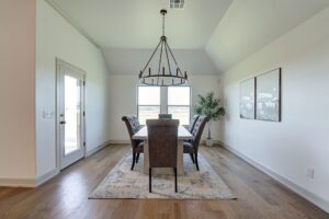 Dining room with a long table, six chairs, and a candle-style chandelier. Large windows, white walls, wooden floor, and a potted plant complete the space.