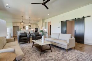 Modern living room with beige and gray seating, a patterned rug, and a white coffee table. Kitchen with pendant lights in the background. Sliding barn doors are visible on the right.