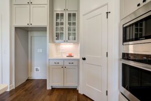 Modern kitchen with white cabinets, glass-paneled doors, built-in oven, and wooden flooring. Small alcove with recessed lighting displays a few decorative items.