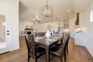 Open-concept dining and living area with a wooden table, chairs, and modern chandelier. Kitchen with white cabinets and pendant lights in the background. Cozy living room with a fireplace.