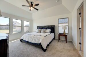 Bedroom with a large bed, ceiling fan, nightstand with a lamp, and three windows allowing natural light. Walls and ceiling are white, and the floor is carpeted.