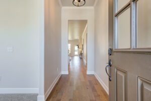 A wooden front door opens into a hallway with hardwood flooring, white walls, and a ceiling light fixture, leading to a bright room at the end.