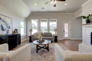 A modern living room with large windows, a beige sofa, two white armchairs, a round wooden coffee table, and a fireplace. Walls are light-colored with abstract art. Ceiling fan above.