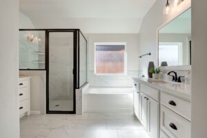 A modern bathroom with a glass shower, a bathtub, a large mirror, and a white vanity with dual sinks. The room features white marble flooring and walls.