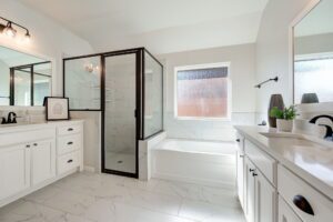 Spacious bathroom with a glass shower, white bathtub, two sinks with mirrors, and tile flooring. Bright light from a window enhances the clean, modern design.