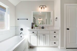 Bright bathroom with a white vanity, large mirror, bathtub, and a window. The room features modern fixtures and neutral tones.