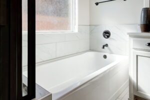 A modern bathroom features a white bathtub with a tiled surround, a dark faucet, and a frosted window above.