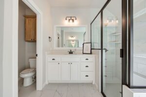 Modern bathroom with a glass shower, white vanity with dual sinks, large mirror, and a toilet. Tiled floor and walls, overhead lighting, and a small wooden cabinet.